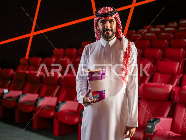 رجل عربي خليجي سعودي في صالة السينما، مبتسم وينظر إلى الكاميرا، يرتدي الزي السعودي التقليدي، يحمل بيده دلو الفشار، أنشطة ترفيهية، قضاء أوقات ممتعة، مشاهدة الأفلام والعروض المسرحية في السينما، سينما ومسرح، خلفية قاعة عرض السينما، صالة العرض