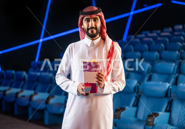 رجل عربي خليجي سعودي في صالة السينما، مبتسم وينظر إلى الكاميرا، يرتدي الزي السعودي التقليدي، يحمل بيده دلو الفشار، أنشطة ترفيهية، قضاء أوقات ممتعة، مشاهدة الأفلام والعروض المسرحية في السينما، سينما ومسرح، خلفية قاعة عرض السينما، صالة العرض