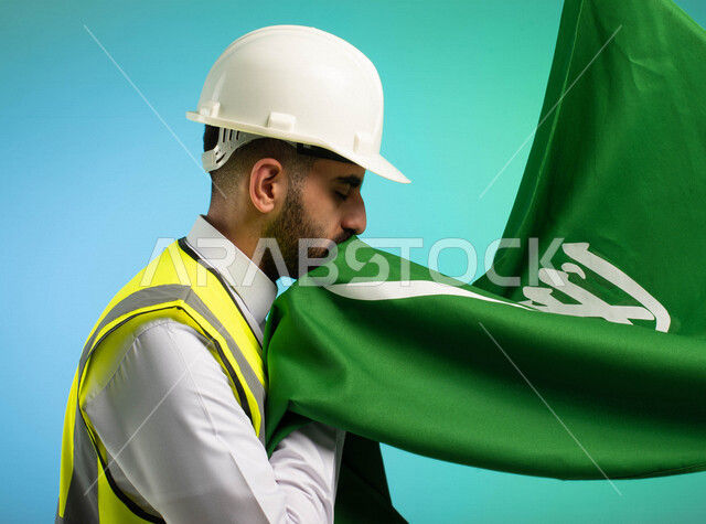 مهندس عربي خليجي سعودي يعمل في مجال الهندسة، يرتدي الخوذة وسترة الحماية، يقبل علم المملكة العربية السعودية، يحتفل باليوم الوطني السعودي، مهن ووظائف السعودية، العمالة السعودية، فرص عمل، مهنة الهندسة، اليوم الوطني السعودي، خلفية كروما
