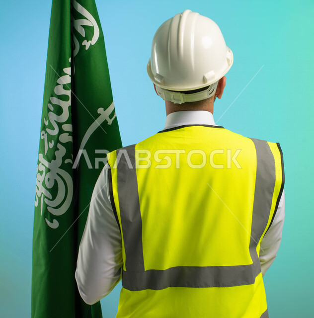 زاوية من الخلف، مهندس عربي خليجي سعودي يعمل في مجال الهندسة، يرتدي الخوذة وسترة الحماية، يحمل علم المملكة العربية السعودية، يحتفل باليوم الوطني، مهن ووظائف السعودية، العمالة السعودية، فرص عمل، مهنة الهندسة، اليوم الوطني السعودي، خلفية كروما