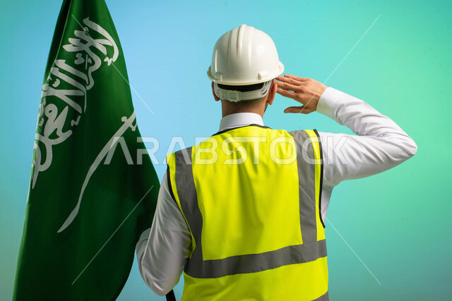 زاوية من الخلف، مهندس عربي خليجي سعودي يعمل في مجال الهندسة، يرتدي الخوذة وسترة الحماية، يحمل علم المملكة و يلقي تحية السلام الوطني، يحتفل باليوم الوطني، مهن ووظائف السعودية، العمالة السعودية، فرص عمل، مهنة الهندسة، اليوم الوطني السعودي، خلفية كروما