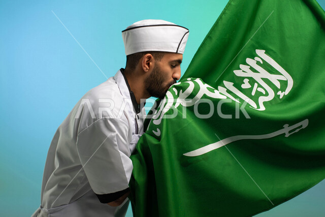 طاه وطباخ عربي خليجي سعودي يعمل في مجال فنون الطهي، يرتدي الزي الرسمي الخاص بمزاولة مهنة الشيف، يقبل علم المملكة العربية السعودية، يحتفل باليوم الوطني، مهن خدمات الأطعمة، العمالة السعودية، فرص عمل، مهنة طاهي الطعام، اليوم الوطني السعودي، خلفية كروما