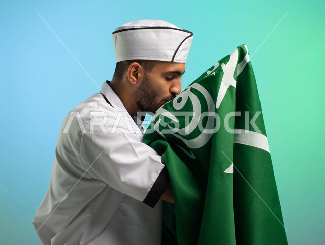 A Saudi Arabian Gulf chef and cook working in the field of culinary arts, wearing the uniform of practicing the profession of chef, accepting the flag of the Kingdom of Saudi Arabia, celebrating the National Day, food service professions, Saudi employment