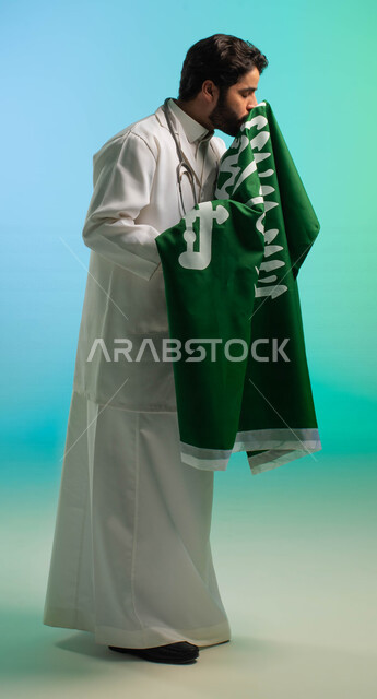 طبيب عربي خليجي سعودي يعمل في قطاع الرعاية الصحية، يرتدي المعطف الطبي، يقبل علم المملكة العربية السعودية، يحتفل باليوم الوطني، مهن ووظائف طبية، العمالة السعودية، فرص عمل، مهنة الطب، اليوم الوطني السعودي، خلفية كروما، صورة بالطول الكامل
