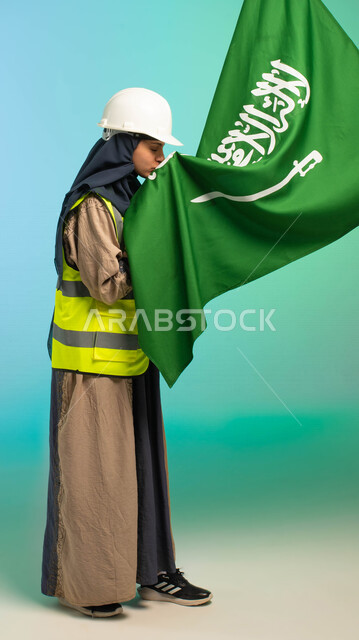 مهندسة عربية خليجية سعودية تعمل في مجال الهندسة، ترتدي الخوذة وسترة الحماية، تقبل علم المملكة العربية السعودية، تحتفل باليوم الوطني، مهن ووظائف السعودية، العمالة السعودية، فرص عمل، مهنة الهندسة، اليوم الوطني السعودي، خلفية كروما، صورة بالطول الكامل