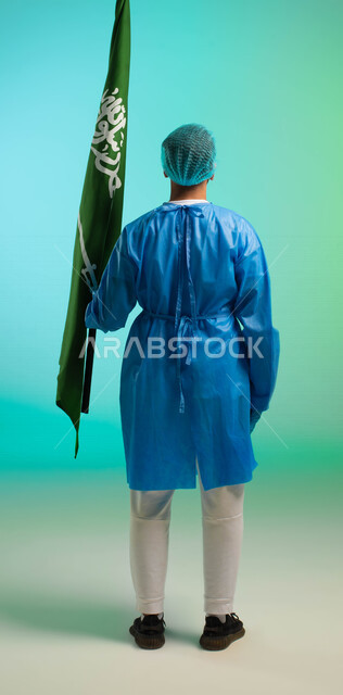 زاوية من الخلف، طبيب جراح عربي خليجي سعودي يعمل في قطاع الرعاية الصحية، يرتدي زي العمليات، يحمل علم السعودية، يحتفل باليوم الوطني، مهن ووظائف طبية، العمالة السعودية، فرص عمل، مهنة الطب، اليوم الوطني السعودي، خلفية كروما، صورة بالطول الكامل