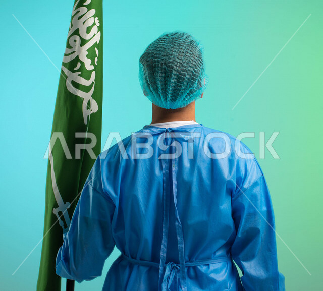 زاوية من الخلف، طبيب جراح عربي خليجي سعودي يعمل في قطاع الرعاية الصحية، يرتدي زي العمليات، يحمل علم المملكة العربية السعودية، يحتفل باليوم الوطني السعودي، مهن ووظائف طبية، العمالة السعودية، فرص عمل، مهنة الطب، اليوم الوطني السعودي، خلفية كروما