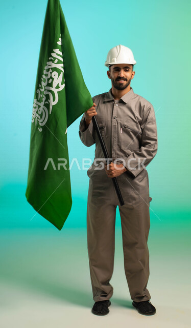 مهندس فني عربي خليجي سعودي يعمل في قطاع الإنشاءات، يرتدي خوذة الحماية، يحمل علم السعودية، مهن ووظائف سعودية، العمالة السعودية، فرص عمل، مهنة فني كهرباء، الأعمال والصيانة، اليوم الوطني السعودي، خلفية كروما، صورة بالطول الكامل