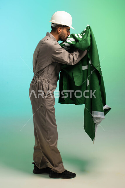 مهندس فني عربي خليجي سعودي يعمل في قطاع الإنشاءات، يرتدي خوذة الحماية، يقبل علم السعودية، مهن ووظائف سعودية، العمالة السعودية، فرص عمل، مهنة فني كهرباء، الأعمال والصيانة، اليوم الوطني السعودي، خلفية كروما، صورة بالطول الكامل