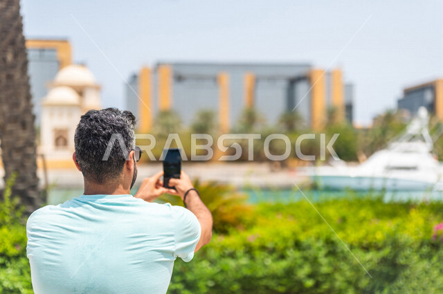 زاوية من الخلف لرجل عربي خليجي سعودي يقضي أوقات ممتعة في كورنيش جدة، إلتقاط صور تذكاريه بإستخدام الهاتف المحمول، موهبة التصوير، واجهة جدة البحرية، أماكن سياحية ترفيهية بالمملكة العربية السعودية، شاطئ سياحي