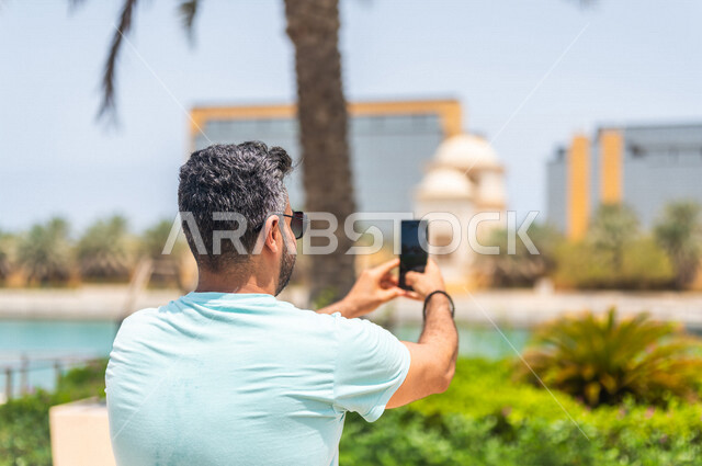 زاوية من الخلف لرجل عربي خليجي سعودي يقضي أوقات ممتعة في كورنيش جدة، إلتقاط صور تذكاريه بإستخدام الهاتف المحمول، موهبة التصوير، واجهة جدة البحرية، أماكن سياحية ترفيهية بالمملكة العربية السعودية، شاطئ سياحي