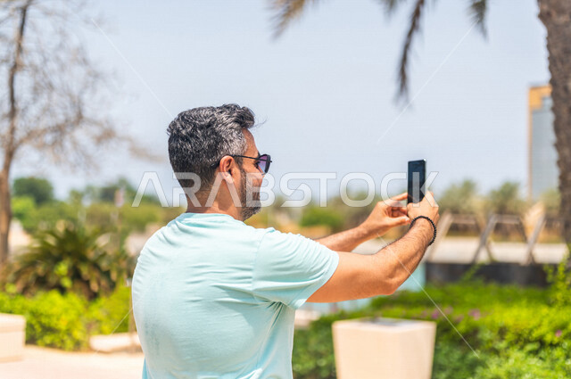 زاوية من الخلف لرجل عربي خليجي سعودي يقضي أوقات ممتعة في كورنيش جدة، إلتقاط صور تذكاريه بإستخدام الهاتف المحمول، موهبة التصوير، واجهة جدة البحرية، أماكن سياحية ترفيهية بالمملكة العربية السعودية، شاطئ سياحي