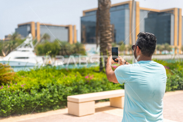 زاوية من الخلف لرجل عربي خليجي سعودي يقضي أوقات ممتعة في كورنيش جدة، إلتقاط صور تذكاريه بإستخدام الهاتف المحمول، موهبة التصوير، واجهة جدة البحرية، أماكن سياحية ترفيهية بالمملكة العربية السعودية، شاطئ سياحي
