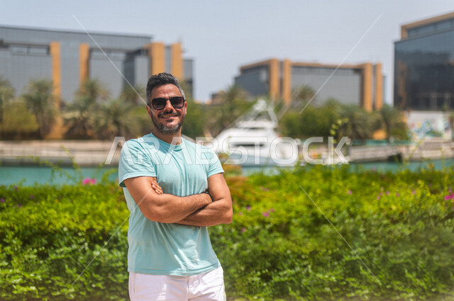 رجل عربي خليجي سعودي مبتسم، يرتدي النظارة الشمسية، يقف بإستقامة بأيدي متقاطعة على كورنيش جدة، كورنيش جدة السياحي، واجهة جدة البحرية، أماكن سياحية ترفيهية بالمملكة العربية السعودية، الإستجمام والتنزه في الهواء الطلق، أنشطة ترفيهية في جدة