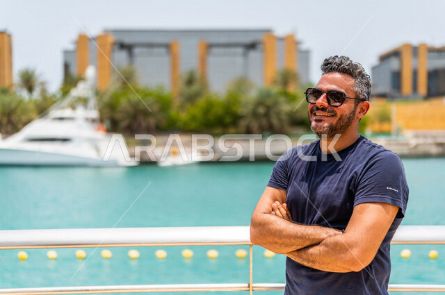 رجل عربي خليجي سعودي مبتسم، يرتدي النظارة الشمسية، يقف بإستقامه بأيدي متقاطعة على كورنيش جدة، كورنيش جدة السياحي، واجهة جدة البحرية، أماكن سياحية ترفيهية بالمملكة العربية السعودية، الإستجمام والتنزه ، خلفية ضبابية، أنشطة ترفيهية في جدة