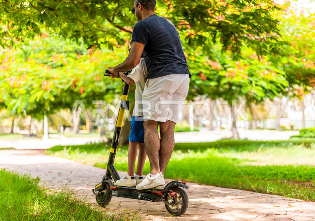 زاوية من الخلف لرجل عربي خليجي سعودي يقضي وقت ممتع بقيادة السكوتر الكهربائي مع إبنه في إحدى الحدائق السياحية الترفيهية في المملكة العربية السعودية، مشاركة الأبناء الأنشطة الترفيهية، قضاء أوقات عائلية ممتعة، مفهوم الأسرة السعيدة، خلفية الطبيعة، حدائق عامة