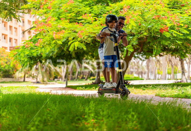 A Saudi Arabian Gulf man spends a fun time driving the electric scooter with his son in one of the recreational tourist parks in the Kingdom of Saudi Arabia, children’s participation in recreational activities, expressions of happiness, spending enjoyable