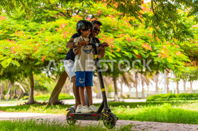 A Saudi Arabian Gulf man spends a fun time driving the electric scooter with his son in one of the recreational tourist parks in the Kingdom of Saudi Arabia, children’s participation in recreational activities, expressions of happiness, spending enjoyable
