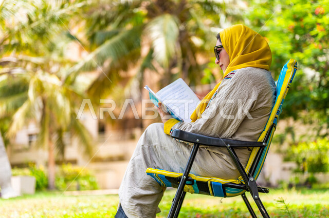 زاوية من الجنب لمرأة عربية خليجية سعودية محجبة، ترتدي العباءة الخليجية ونظارة شمسية، تحمل بيدها كتاب وتجلس في إحدى الحدائق السياحية الترفيهية في المملكة العربية السعودية، قراءة الكتب، جلسات هادئة، خلفية الطبيعة، حدائق عامة