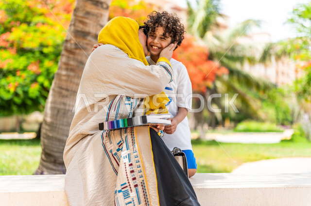 مرأة عربية خليجية سعودية تقضي وقت ممتع برفقة إبنها في إحدى الحدائق السياحية الترفيهية في المملكة العربية السعودية، تقبل إبنها بتعابير تدل على السعادة، تحمل بيدها كتاب، قضاء أوقات عائلية ممتعة، مفهوم الأسرة السعيدة، جلسات هادئة، خلفية الطبيعة، حدائق عامة