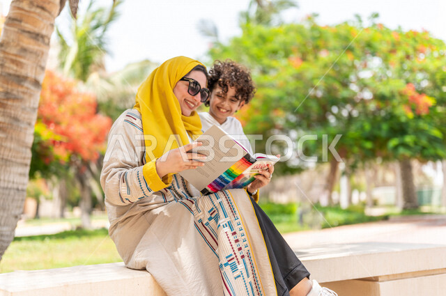 مرأة عربية خليجية سعودية مع إبنها يقضيان وقت ممتع في إحدى الحدائق السياحية الترفيهية في المملكة العربية السعودية، مبتسمان وينظران إلى الكتاب، مفهوم الأسرة السعيدة، مشاركة الأبناء بقراءة القصص والروايات، جلسات هادئة، خلفية الطبيعة، حدائق عامة