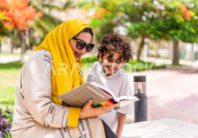 مرأة عربية خليجية سعودية مع إبنها يقضيان وقت ممتع في إحدى الحدائق السياحية الترفيهية في المملكة العربية السعودية، مبتسمان وينظران إلى الكتاب، مفهوم الأسرة السعيدة، مشاركة الأبناء بقراءة القصص والروايات، جلسات هادئة، خلفية الطبيعة، حدائق عامة