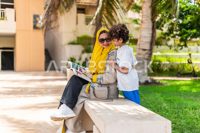 مرأة عربية خليجية سعودية مع إبنها يقضيان وقت ممتع في إحدى الحدائق السياحية الترفيهية في المملكة العربية السعودية، مبتسمان وينظران إلى الكتاب، مفهوم الأسرة السعيدة، مشاركة الأبناء بقراءة القصص والروايات، جلسات هادئة، خلفية الطبيعة، حدائق عامة