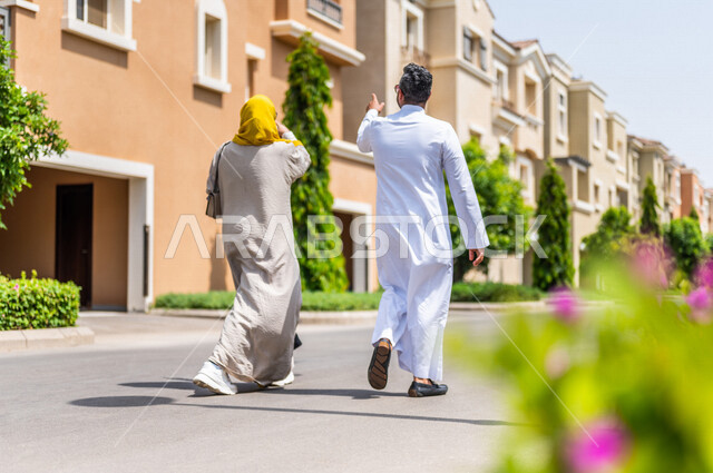 زاوية من الخلف لزوجان عربيان خليجيان سعوديان يتجولان في الأحياء السكنية الراقية بالسعودية، ينظران إلى العمارات السكنية، كمباوند سكني، مجمع سكني، الإسكان العائلي بالسعودية، تصاميم هندسية معمارية حديثة، أسرة سعودية، جودة الحياة