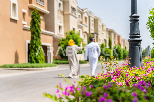 زاوية من الخلف لزوجان عربيان خليجيان سعوديان يتجولان في الأحياء السكنية الراقية بالسعودية، ينظران إلى العمارات السكنية، كمباوند سكني، مجمع سكني، الإسكان العائلي بالسعودية، تصاميم هندسية معمارية حديثة، أسرة سعودية، جودة الحياة