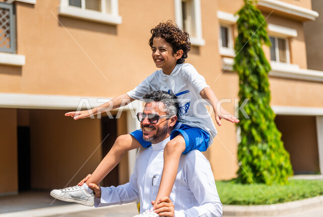 عائلة عربية خليجية سعودية تقضي أوقات ممتعة في الأحياء السكنية الراقية بالسعودية، كمباوند سكني، فلل سكنية، مجمع سكني، الإسكان العائلي في المملكة العربية السعودية، أجواء عائلية سعيدة، الأب يحمل إبنه على كتفيه، الابن سعيد باللعب، أسرة سعودية، جودة الحياة