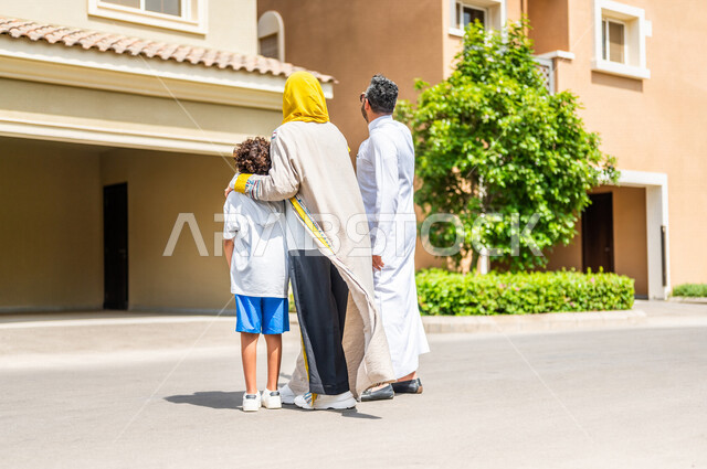زاوية من الخلف لعائلة عربية خليجية سعودية تتجول في الأحياء السكنية الراقية بالسعودية، ينظران إلى الفيلا السكنية، كمباوند سكني، فلل سكنية، مجمع سكني، الإسكان العائلي في السعودية، أجواء عائلية سعيدة، أسرة سعودية، جودة الحياة
