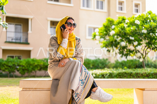 صورة مقربة لمرأة عربية خليجية سعودية محجبة، ترتدي العباءة الخليجية ونظارة شمسية، تقوم بإجراء مكالمة هاتفية بإستخدام الهاتف المحمول في الهواء الطلق في الأحياء السكنية الراقية بالسعودية، كمباوند سكني، الإتصال والدردشة، فلل سكنية بالسعودية، جودة الحياة