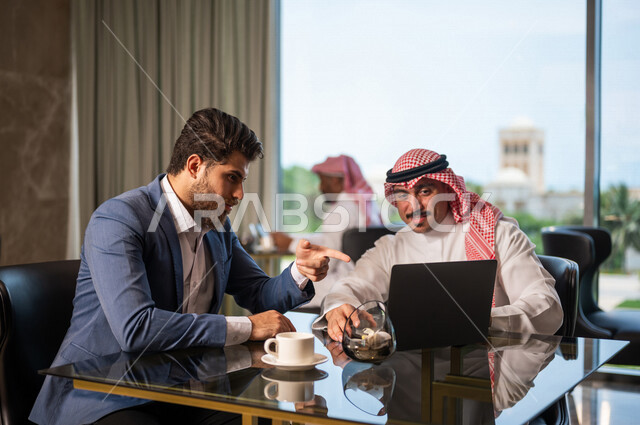 رجل أعمال عربي سعودي خليجي مع رجل أعمال عربي يرتدي البدلة الرسمية, الجلوس في المقهى، إستخدام جهاز الحاسوب المحمول، مناقشة مهام العمل، شركة سعودية، بيئة عمل سعودي، زملاء عمل، الشراكة والتعاون، إجتماع عمل, متابعة الأعمال, تحقيق الأهداف 
