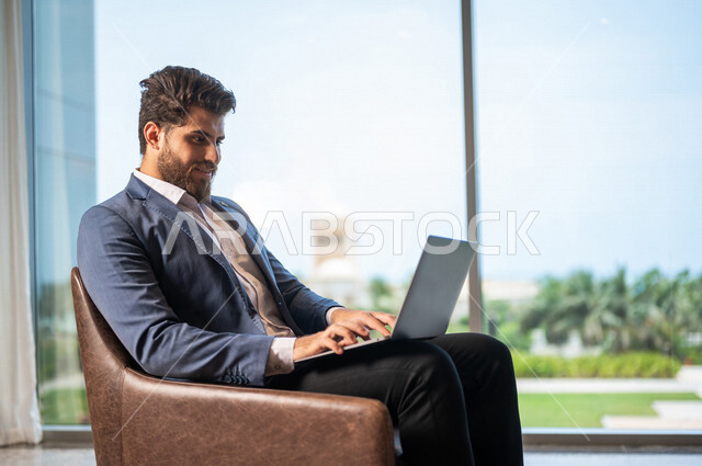 رجل اعمال عربي يرتدي البدلة الرسميه يجلس على كرسي، إستخدام جهاز الحاسوب المحمول, ادارة العمل عن بعد، إنجاز مهام العمل, التسوق الإلكتروني, غرفة الانتظار، تواصل إجتماعي, إجتماع عمل عن بعد, إجراء مكالمات فيديو, إستغلال وقت الإنتظار