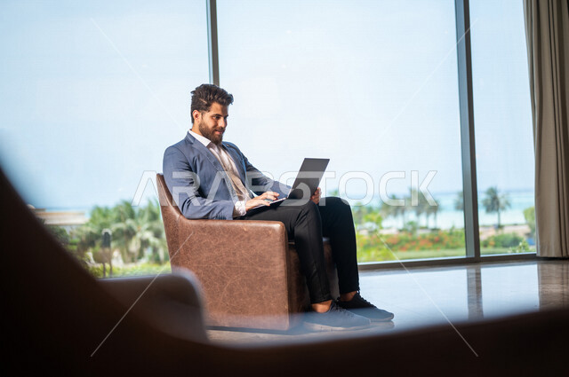 رجل اعمال عربي يرتدي البدلة الرسميه يجلس على كرسي، إستخدام جهاز الحاسوب المحمول, ادارة العمل عن بعد، إنجاز مهام العمل, التسوق الإلكتروني, غرفة الانتظار، تواصل إجتماعي, إجتماع عمل عن بعد, إجراء مكالمات فيديو, إستغلال وقت الإنتظار