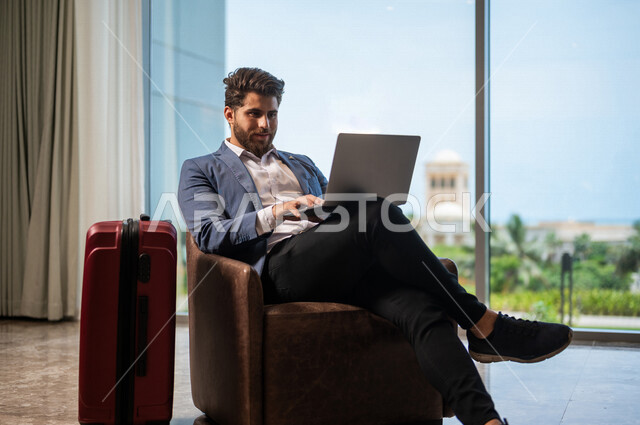 رجل اعمال عربي يرتدي البدلة الرسميه يجلس على كرسي، وبجانبه حقيبة السفر, إستخدام جهاز الحاسوب المحمول, ادارة العمل عن بعد، إنجاز مهام العمل, التسوق الإلكتروني, الاستعداد للسفر في رحلة عمل, غرفة الانتظار، سياحة وسفر, رحله سياحيه