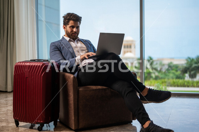 رجل اعمال عربي يرتدي البدلة الرسميه يجلس على كرسي، وبجانبه حقيبة السفر, إستخدام جهاز الحاسوب المحمول, ادارة العمل عن بعد، إنجاز مهام العمل, التسوق الإلكتروني, الاستعداد للسفر في رحلة عمل, غرفة الانتظار، سياحة وسفر, رحله سياحيه