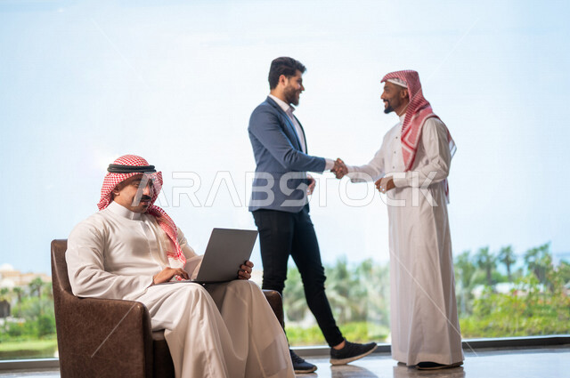 رجل أعمال عربي سعودي خليجي يرتدي الزي السعودي يصافح رجل أعمال أجنبي يرتدي البدلة الرسمية، الإتفاق على المشاريع التجارية، صفقة عمل جديده, شركاء عمل، التعاون بين الشركات، بيئة عمل سعوديه، إتفاقية عمل, شركات عربيه سعوديه خليجيه في المملكه العربيه السعوديه