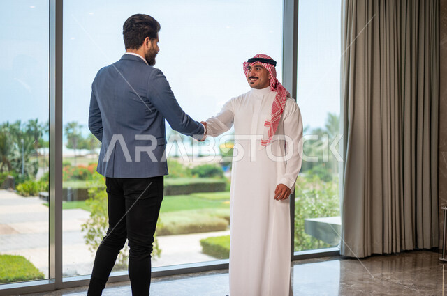 رجل أعمال عربي سعودي خليجي يرتدي الزي السعودي يصافح رجل أعمال عربي يرتدي البدلة الرسمية، الإتفاق على المشاريع التجارية، صفقة عمل جديده, شركاء عمل، التعاون بين الشركات، بيئة عمل سعوديه، إتفاقية عمل, شركات عربيه سعوديه خليجيه في المملكه العربيه السعوديه