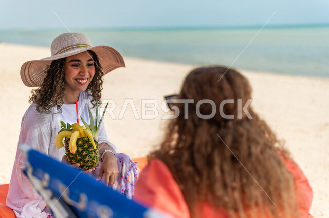 صورة مقربة لصديقتان عربيتان خليجيتان سعوديتان تستمتعان بالاجازة الصيفية على شاطئ بحر جدة، دردشة ومحادثات، الاستمتاع بالمشروبات الصيفية المنعشة على شاطئ البحر، الاسترخاء والترفيه، المتعة والاستجمام على بحر جدة السياحي، ايماءات وجه مبتسمة، ارتداء أزياء صيفي