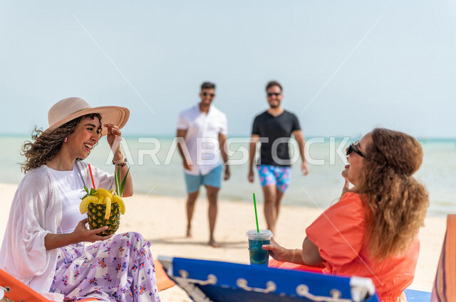 صديقتان عربيتان خليجيتان سعوديتان تستمتعان بالاجازة الصيفية على شاطئ بحر جدة، دردشة ومحادثات، الاستمتاع بالمشروبات الصيفية المنعشة على شاطئ البحر، الاسترخاء والترفيه، المتعة والاستجمام على بحر جدة السياحي، ايماءات وجه مبتسمة، ارتداء أزياء صيفية