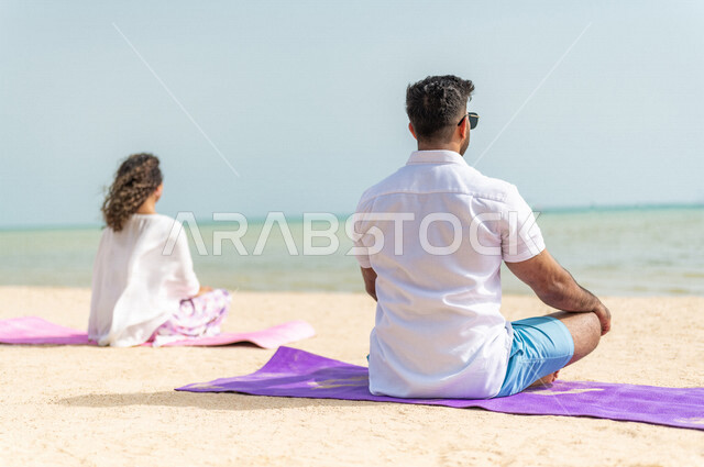 صورة من الخلف لرجل وامرأة عربيان خليجيان سعوديان يمارسان رياضة اليوغا التأملية على شاطيء بحر جدة، ممارسة نشاط رياضي، أنشطة و تمارين رياضية، التأمل والاسترخاء، الرياضة والنشاط البدني، تمارين اللياقة البدنية، أسلوب حياة صحي، لياقة ورشاقة