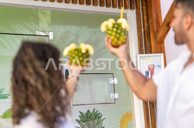 زوجان عربيان خليجيان سعوديان يرفعان حبة الاناناس للاعلى داخل احدى الكافيهات على بحر جدة السياحي، فاكهة الاناناس اللذيذة، ديكورات وتصاميم مميزة، مشروب صيفي منعش، الاستمتاع بالاجازة الصيفية، سياحة وإستجمام، قضاء أوقات ممتعة بالعطلة الصيفية