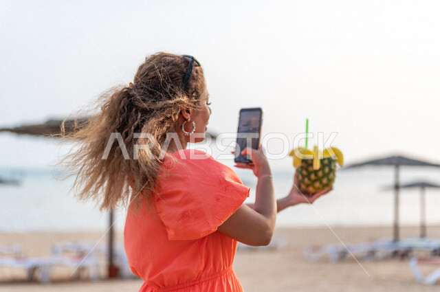 امرأة عربية خليجية سعودية تقوم بتصوير مشروب الاناناس الصيفي، فاكهة الاناناس اللذيذة، شاطئ بحر جدة السياحي، مشروب صيفي منعش، الاستمتاع بالاجازة الصيفية، سياحة وإستجمام، قضاء أوقات ممتعة بالعطلة الصيفية