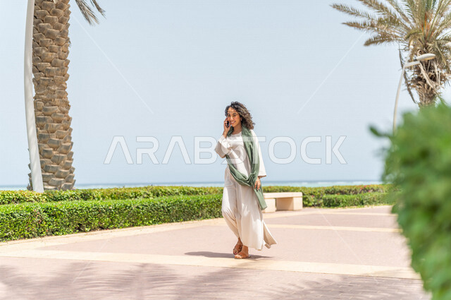 A Saudi Arabian Gulf woman practicing walking in the Jeddah Corniche promenade, making a phone call via a mobile phone, chatting and conversations, a tourist beach, physical activity outdoors, using a mobile phone, communicating with friends, hiking in Je