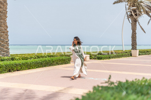 A Saudi Arabian Gulf woman practicing walking in the Jeddah Corniche promenade, making a phone call via a mobile phone, chatting and conversations, a tourist beach, physical activity outdoors, using a mobile phone, communicating with friends, hiking in Je
