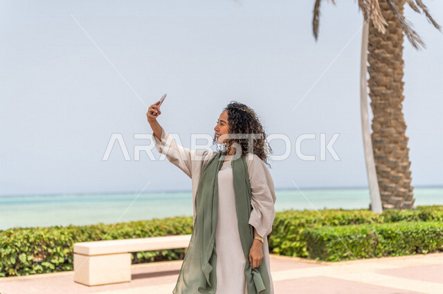 إمرأه عربيه سعوديه خليجيه تقوم بإلتقاط صور تذكاريه في ممشى كورنيش جده, تصوير سلفي, إستخدام الهاتف المحمول, شاطيء سياحي, التنزه في جده, الواجهه البحريه لمدينة جده