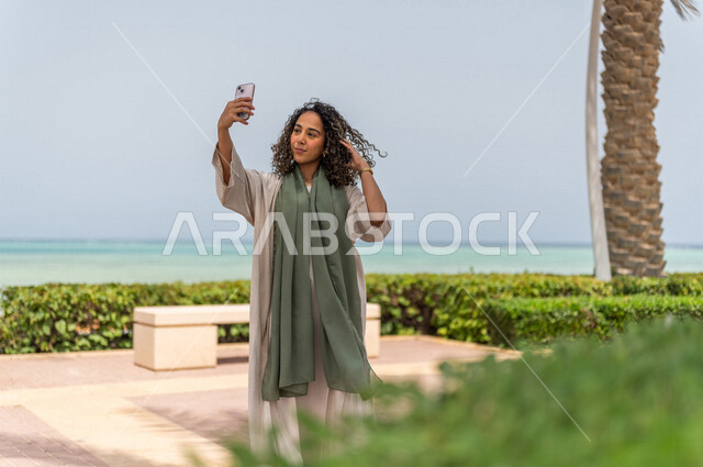 A Saudi Arabian Gulf woman taking souvenir photos on the Jeddah Corniche walkway, taking selfies, using a mobile phone, a tourist beach, hiking in Jeddah, the waterfront of Jeddah