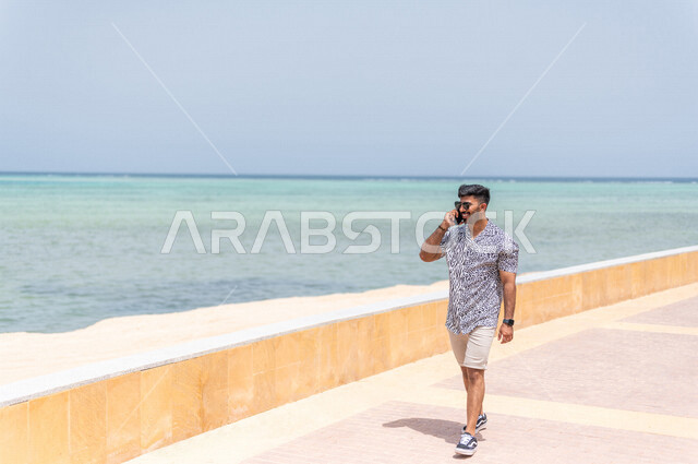 رجل عربي سعودي خليجي يمارس رياضة المشي في ممشى كورنيش جده, إجراء مكالمه هاتفيه عبر الهاتف المحمول, دردشة ومحادثات, التواصل مع الأصدقاء, شاطيء سياحي, التنزه في جده, الواجهه البحريه لمدينة جده