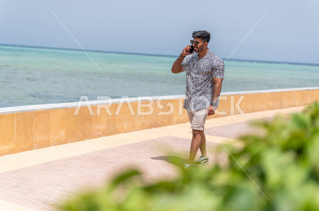 رجل عربي سعودي خليجي يمارس رياضة المشي في ممشى كورنيش جده, إجراء مكالمه هاتفيه عبر الهاتف المحمول, دردشة ومحادثات, التواصل مع الأصدقاء, شاطيء سياحي, التنزه في جده, الواجهه البحريه لمدينة جده